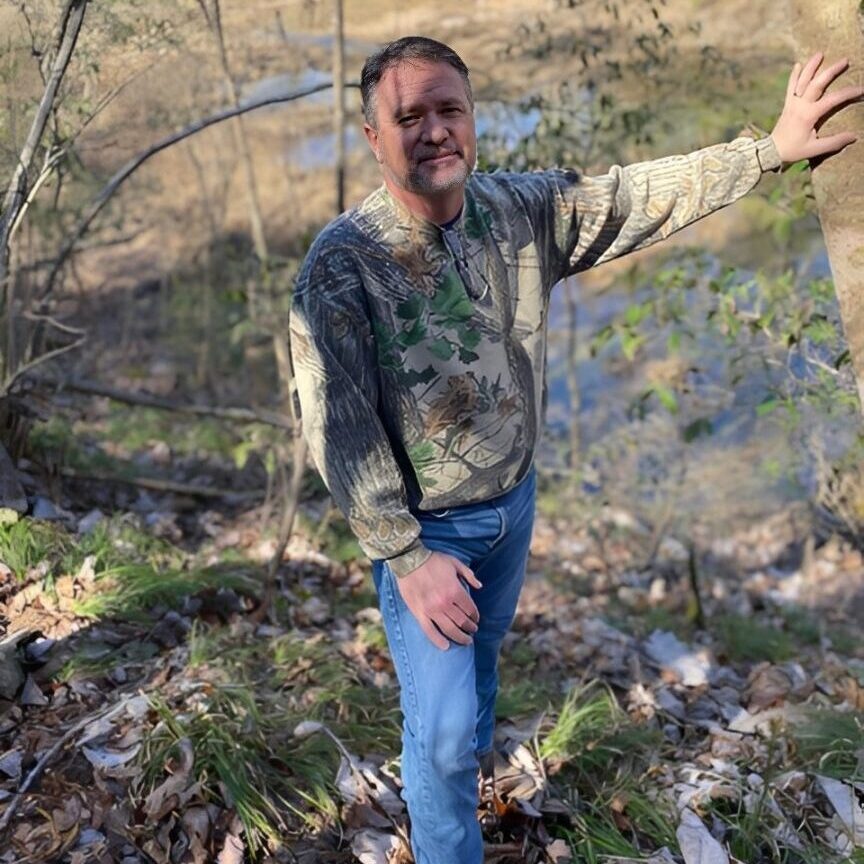 Man smiling outdoors in a wooded area during fall.